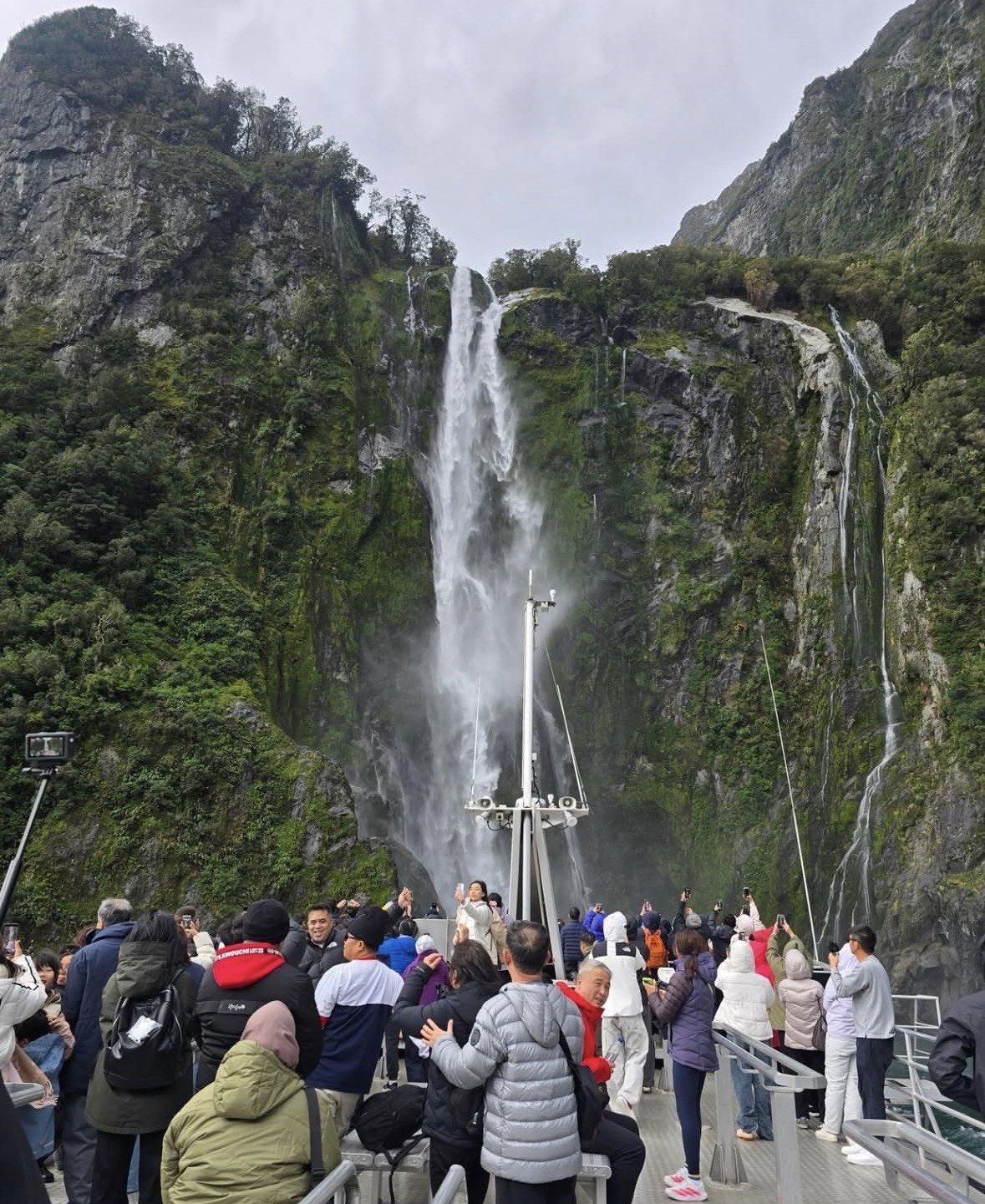 Passengers crowd the bow as a tall waterfall soaks Milford Sound cliffs