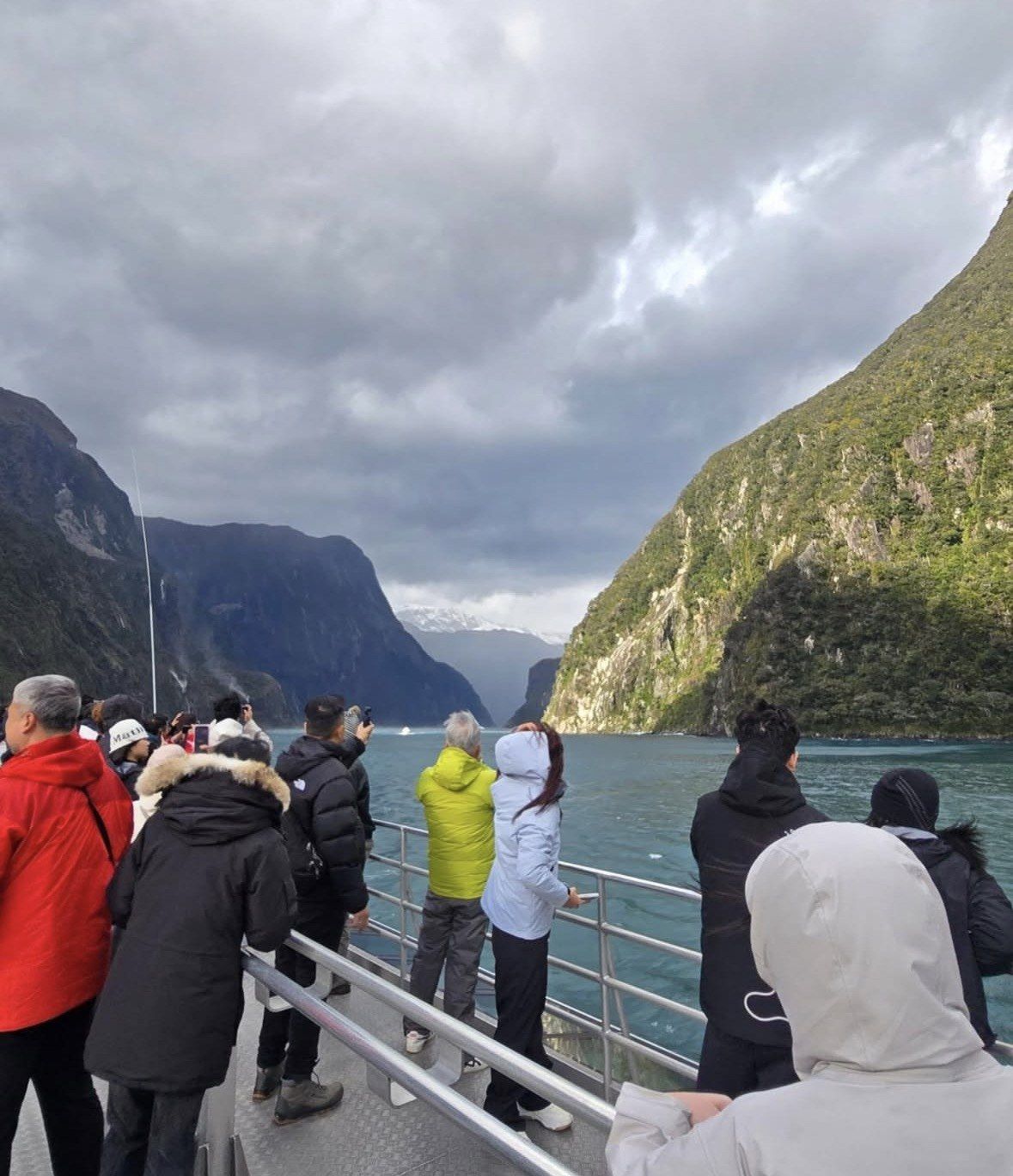 Travellers in winter jackets watch Milford Sound cliffs under storm clouds